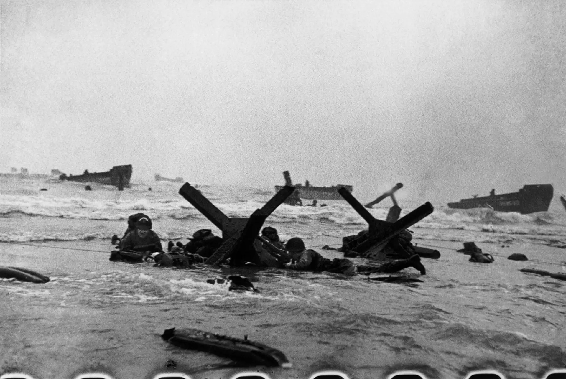 Myndin er tikin tíðliga á morgni 6. juni. Veðrið er enn eitt sindur keðiligt, tó betri enn tað hevði verið kvøldið fyri. Amerikanskir hermenn verja seg aftan fyri forðingar, sum týskarar høvdu lagt at forða landgongubátunum, sum síggjast aftanfyri. Mynd: Robert Capa