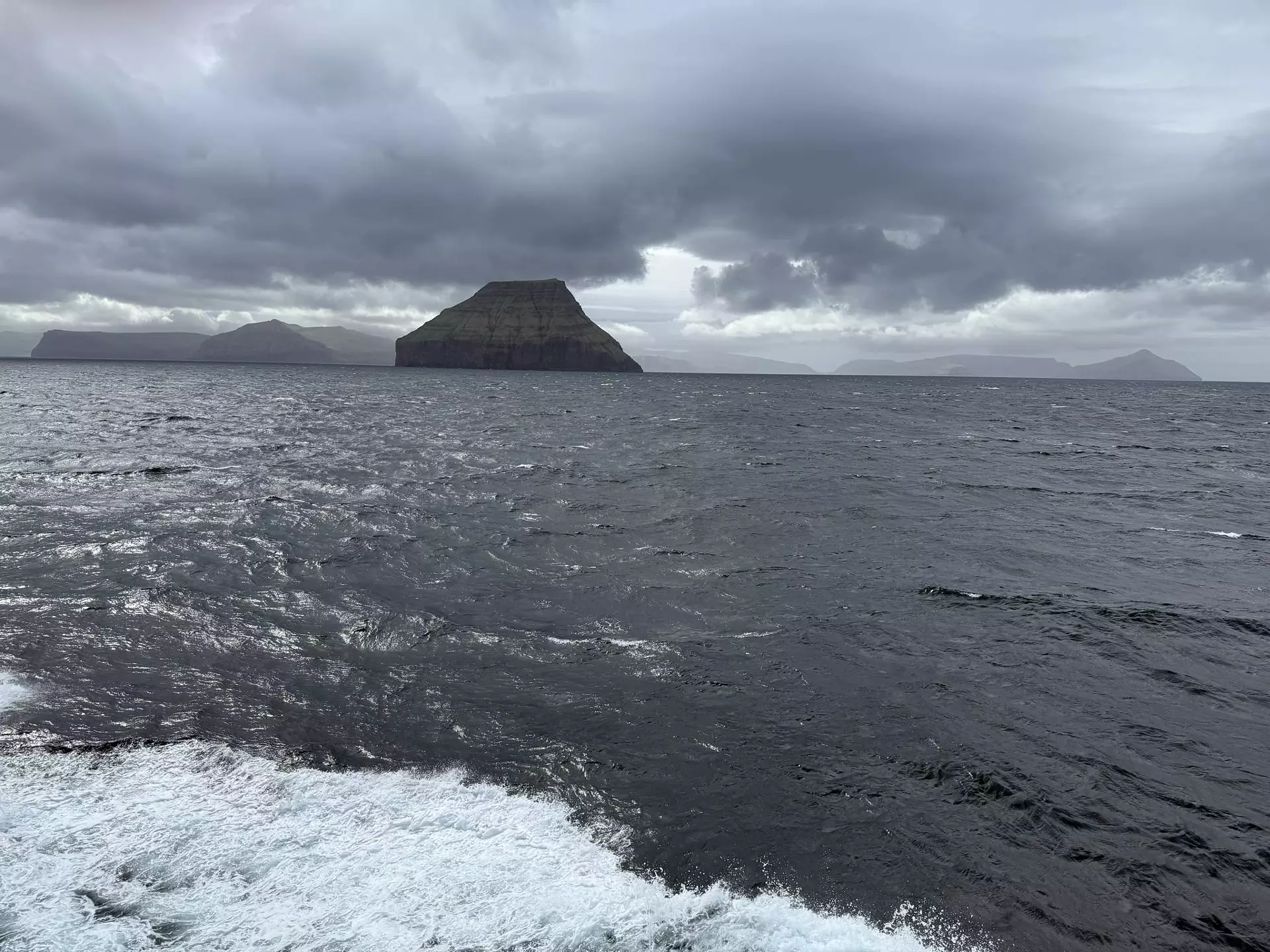 Eitt brúgvalag við regni er yvir Føroyum. Tung skýggj við avfalli (nimbostratus) síggjast yvir øllum oyggjunum á myndini, sum er tikin í Suðuroyarfirði umborð á Smyrli. Lágtrýstið, sum brúgvalagið hoyrdi til, var beint norðan fyri Ísland. Mynd: Finn Johannessen.